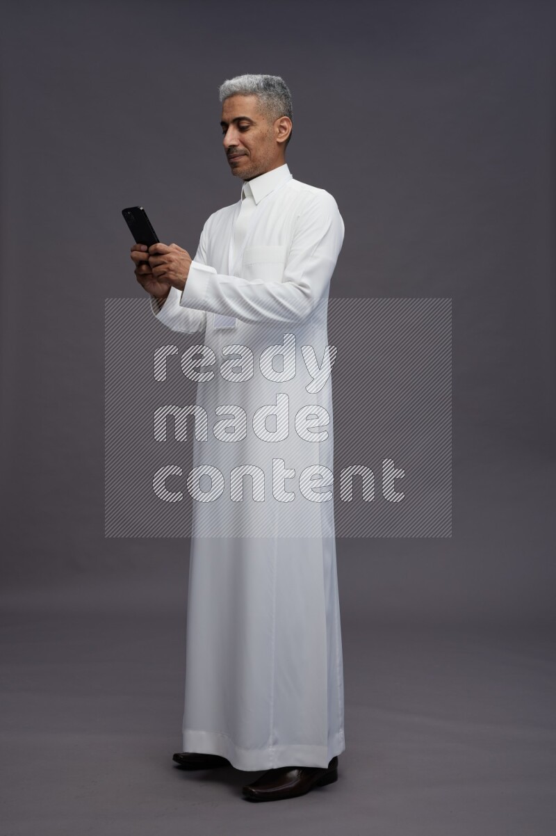 Saudi man wearing thob with neck strap employee badge standing texting on phone on gray background