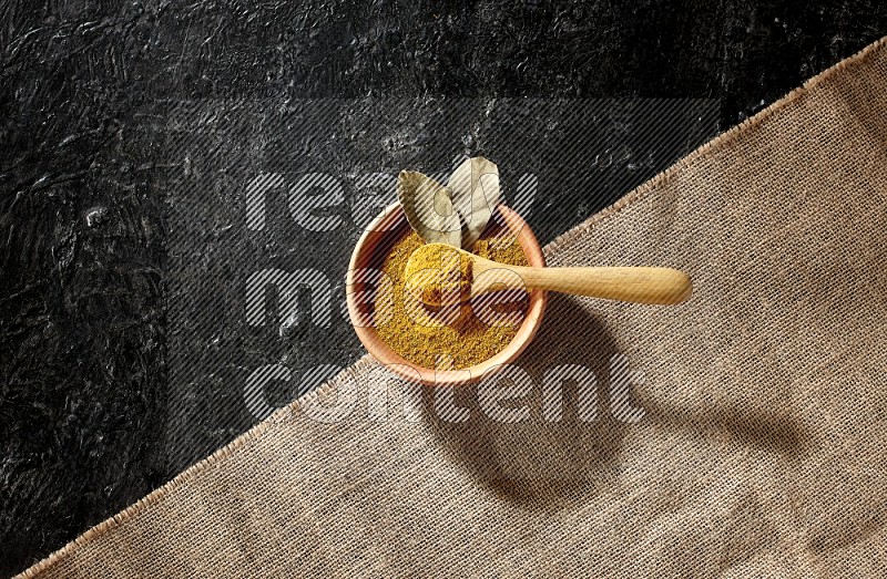 وعاء خشبي وملعقة خشبية ممتلئان ببودرة الكركم على قماش الخيش موضوعان على خلفية سوداء