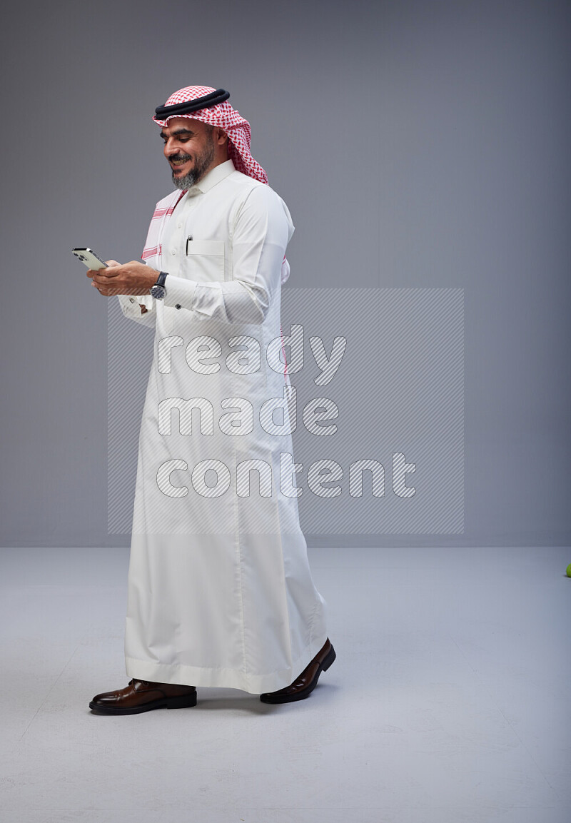 Saudi man Wearing Thob and red Shomag standing texting on phone on Gray background