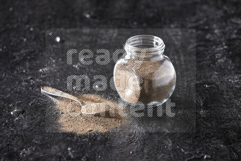 A glass spice jar full of black pepper powder and a metal spoon full of powder on textured black flooring