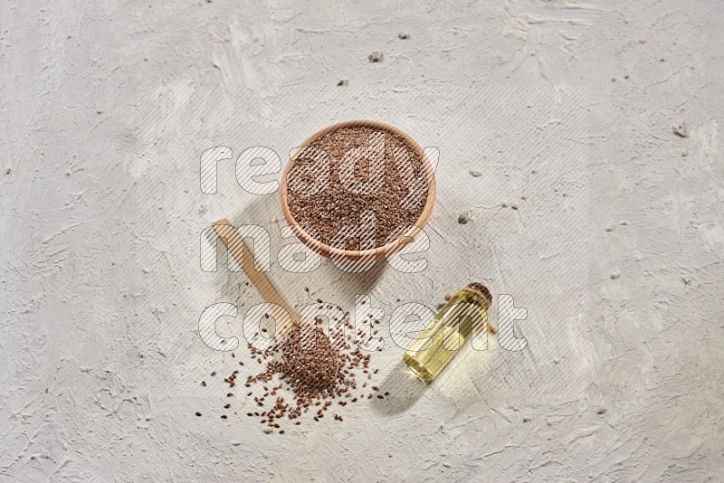 A wooden bowl and spoon full of flaxseeds with a bottle of flaxseeds oil on a textured white flooring