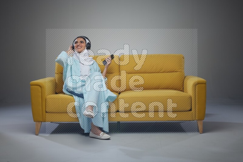A Saudi woman wearing a light blue Abaya and a white head scarf sitting on a yellow sofa and holding an electronic while wearing headphone eye level on a grey background