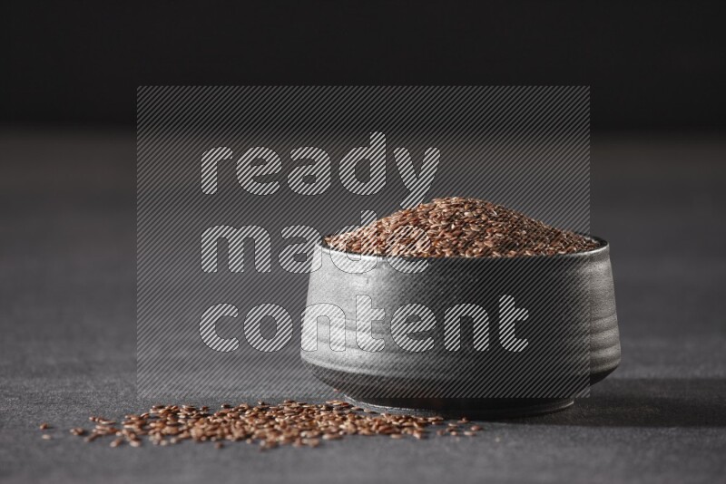 A black pottery bowl full of flaxseeds surrounded by the seeds on a black flooring