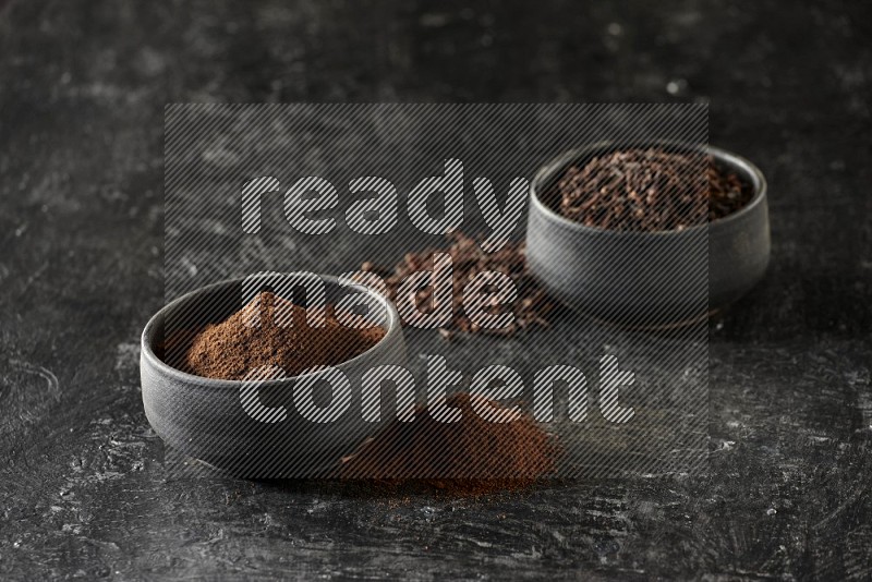 2 Black pottery bowls full of cloves and the other full of cloves powder on textured black flooring