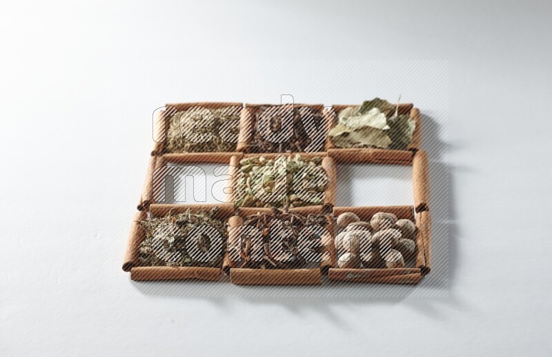 9 squares of cinnamon sticks full of cardamom in the middle surrounded by nutmeg, cinnamon, bay laurel leaves, cloves, cumin, dried ginger, dried basil and star anise on white flooring