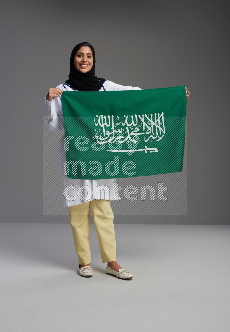 Saudi woman wearing lab coat with stethoscope standing holding Saudi flag on Gray background