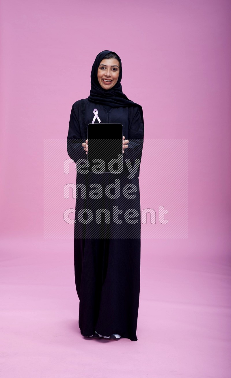 Saudi woman wearing pink ribbon on Abaya standing showing tablet to camera on pink background