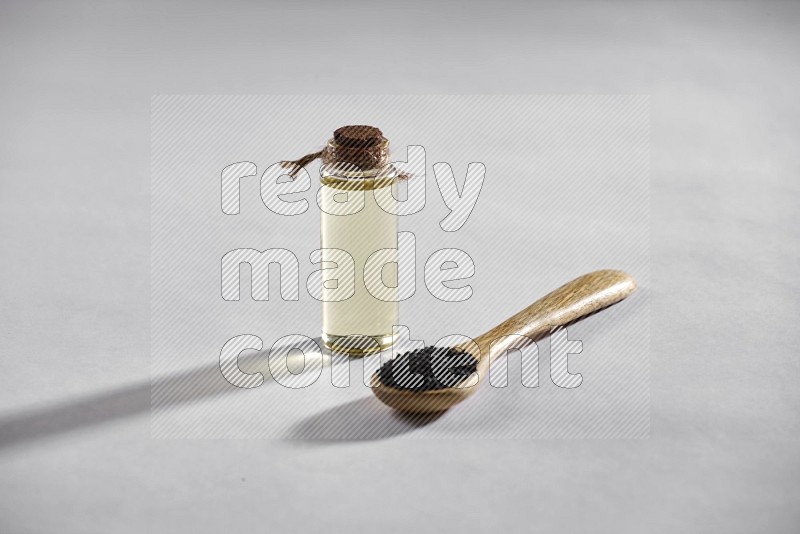 A wooden spoon full of black seeds and a glass bottle of black seeds oil on a white flooring in different angles