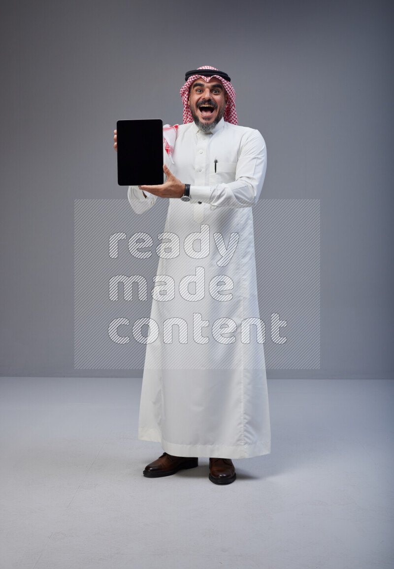 Saudi man Wearing Thob and red Shomag standing showing tablet to camera on Gray background