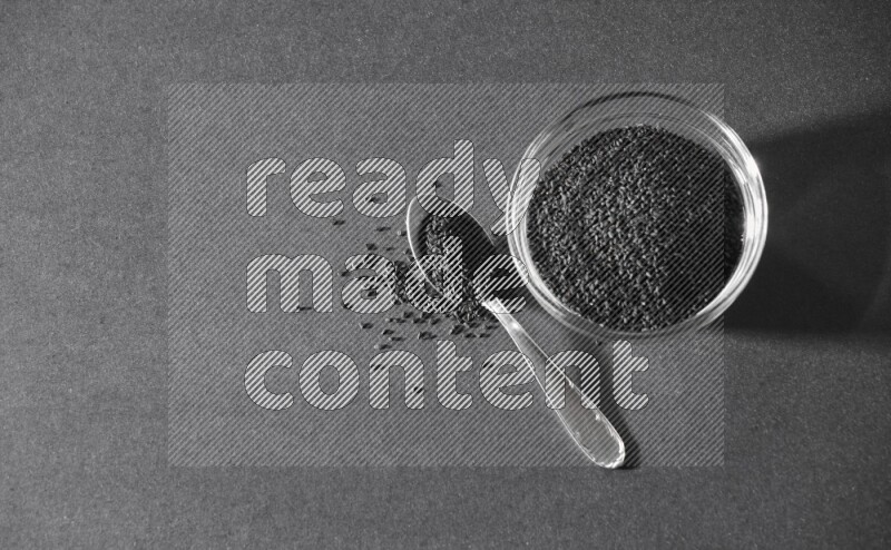 A glass bowl full of black seeds with a metal spoon full of the seeds on a black flooring