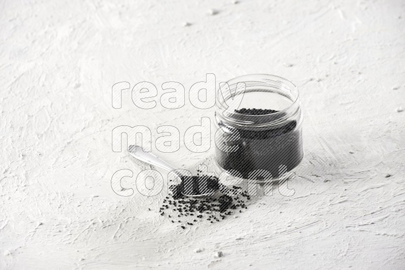 A glass jar and a metal spoon full of black seeds on a textured white flooring in different angles