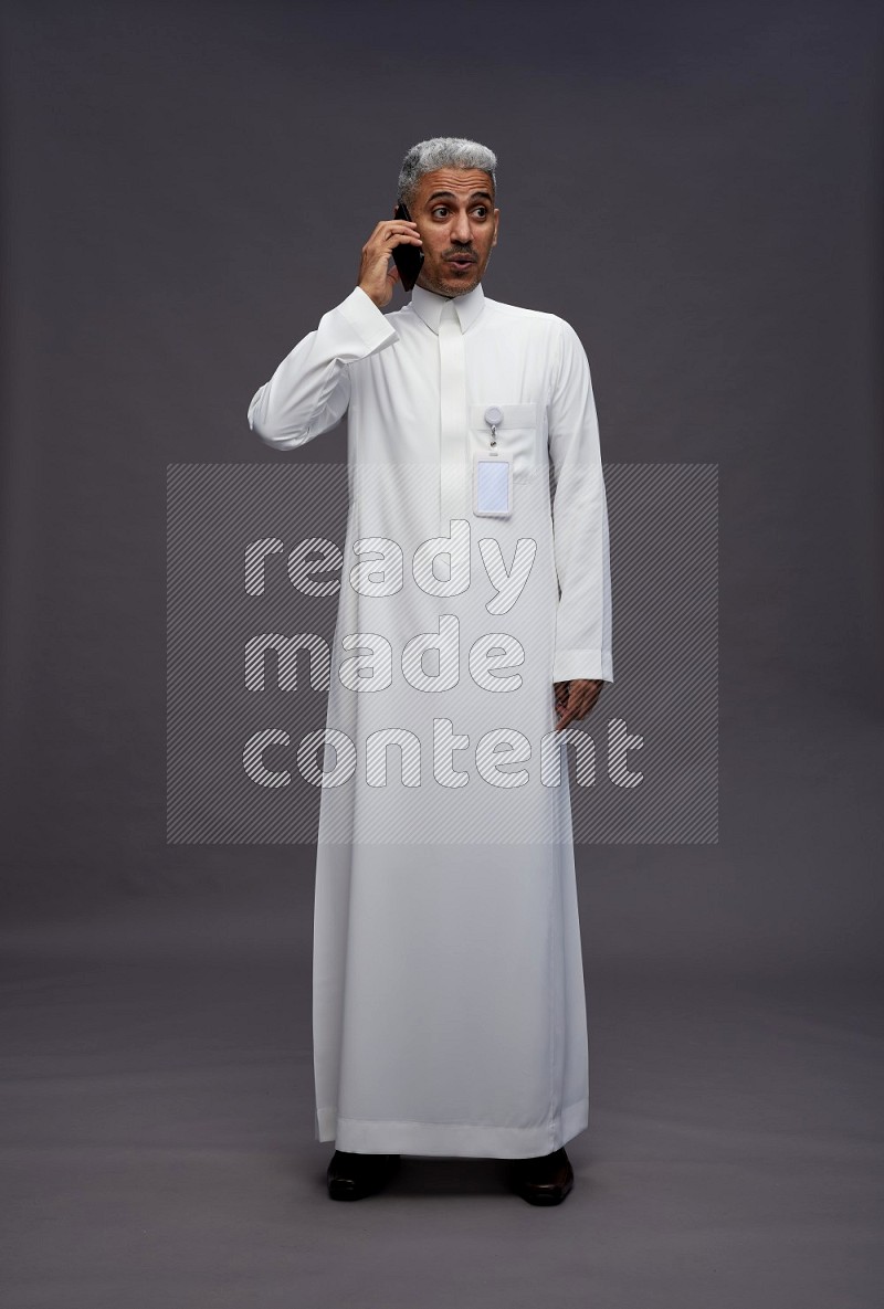 Saudi man wearing thob with pocket employee badge standing talking on phone on gray background
