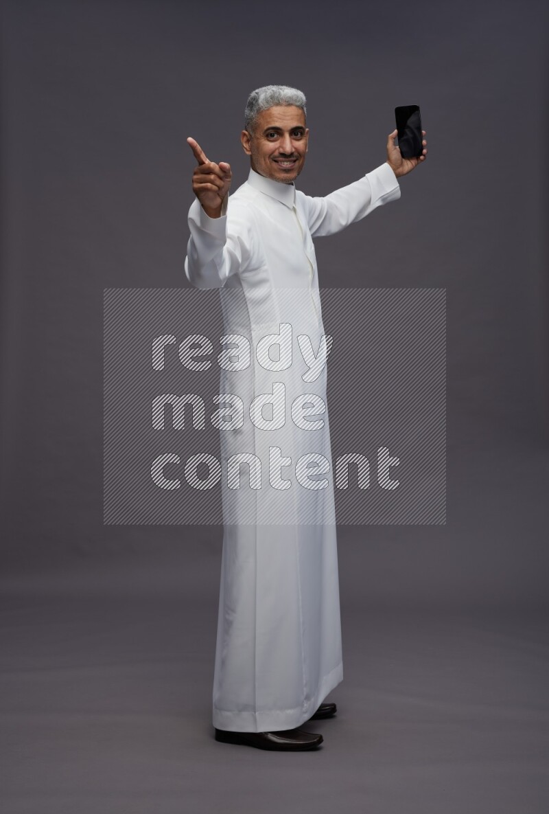 Saudi man wearing thob standing taking selfie on gray background