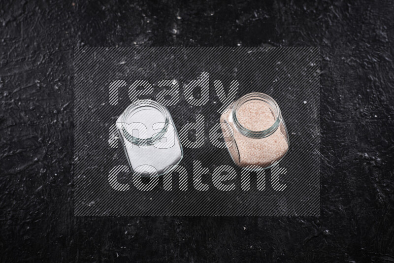 2 glass jars one is filled with fine himalayan salt and the other with table salt on black background