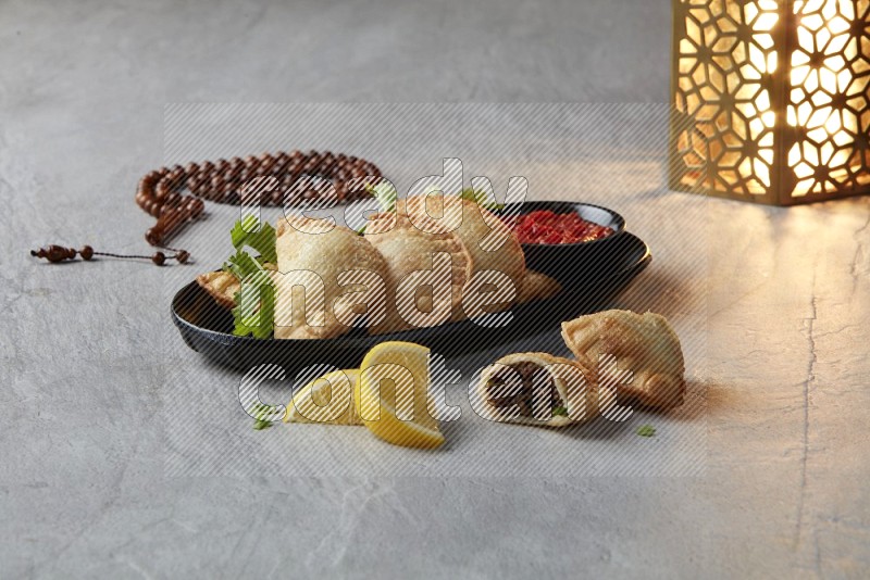 Five fried sambosas in an oval shaped black plate, beside a cut meat sambosa, a brown misbaha and a golden lantern on a gray background