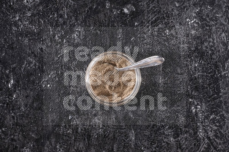 A glass jar full of black pepper powder and a metal spoon on a textured black flooring