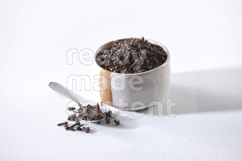 A beige ceramic bowl full of cloves and a metal spoon next to it on a white flooring