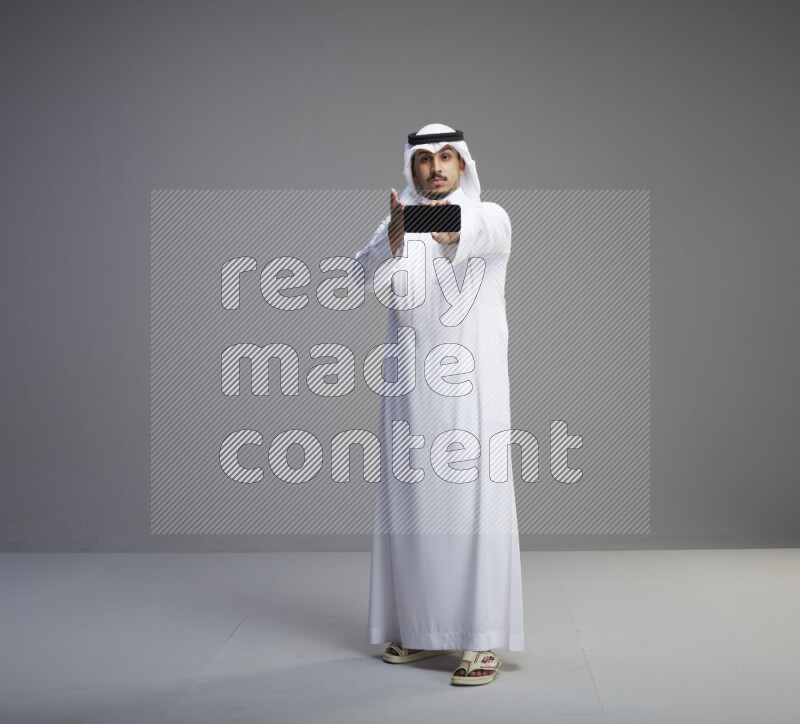 A Saudi man standing wearing thob and white shomag showing phone to camera on gray background