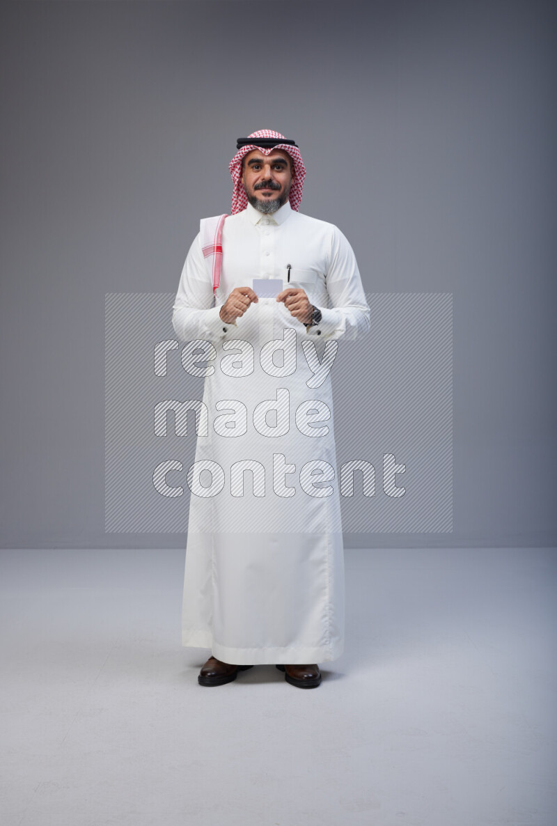 Saudi man Wearing Thob and red Shomag standing holding ATM card on Gray background