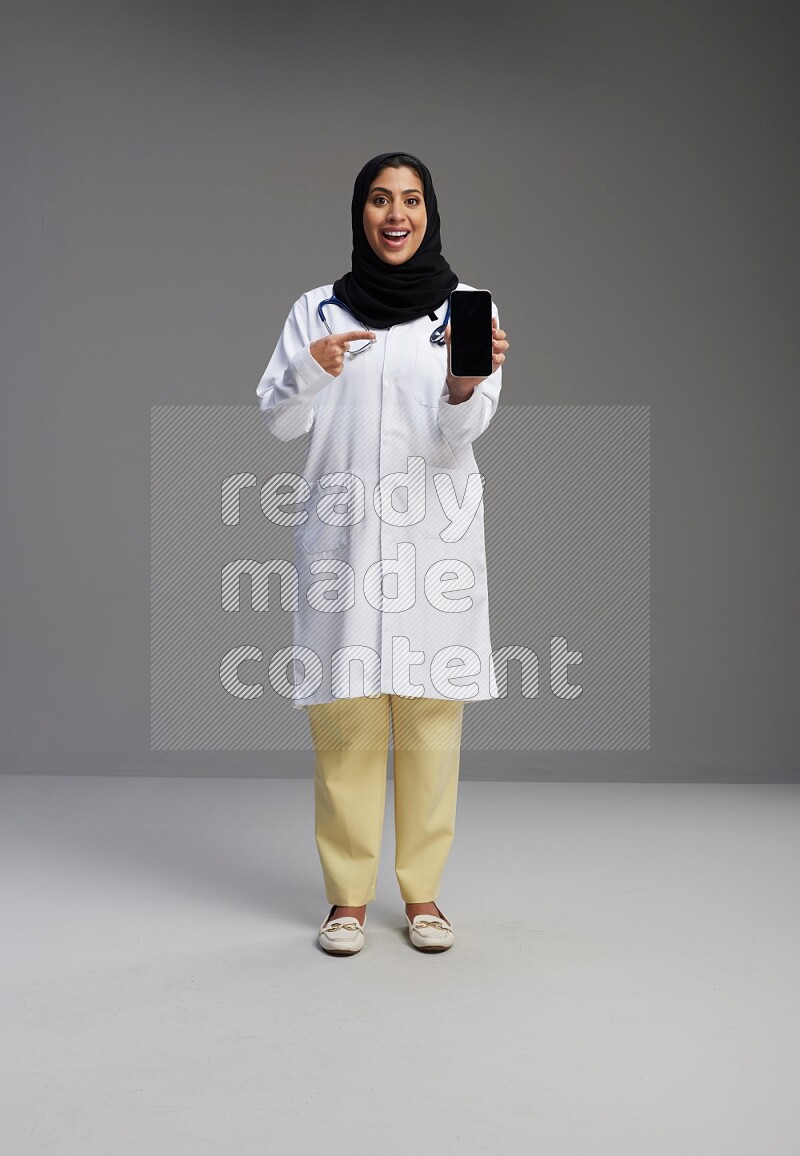 Saudi woman wearing lab coat with stethoscope standing showing phone to camera with sign in the back on Gray background