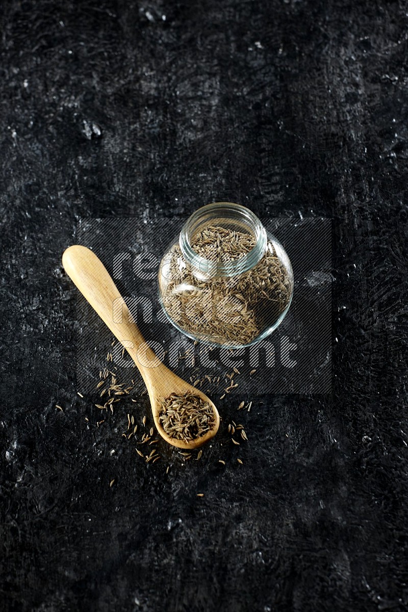 A glass spice jar and a wooden spoon full of cumin seeds on a textured black flooring