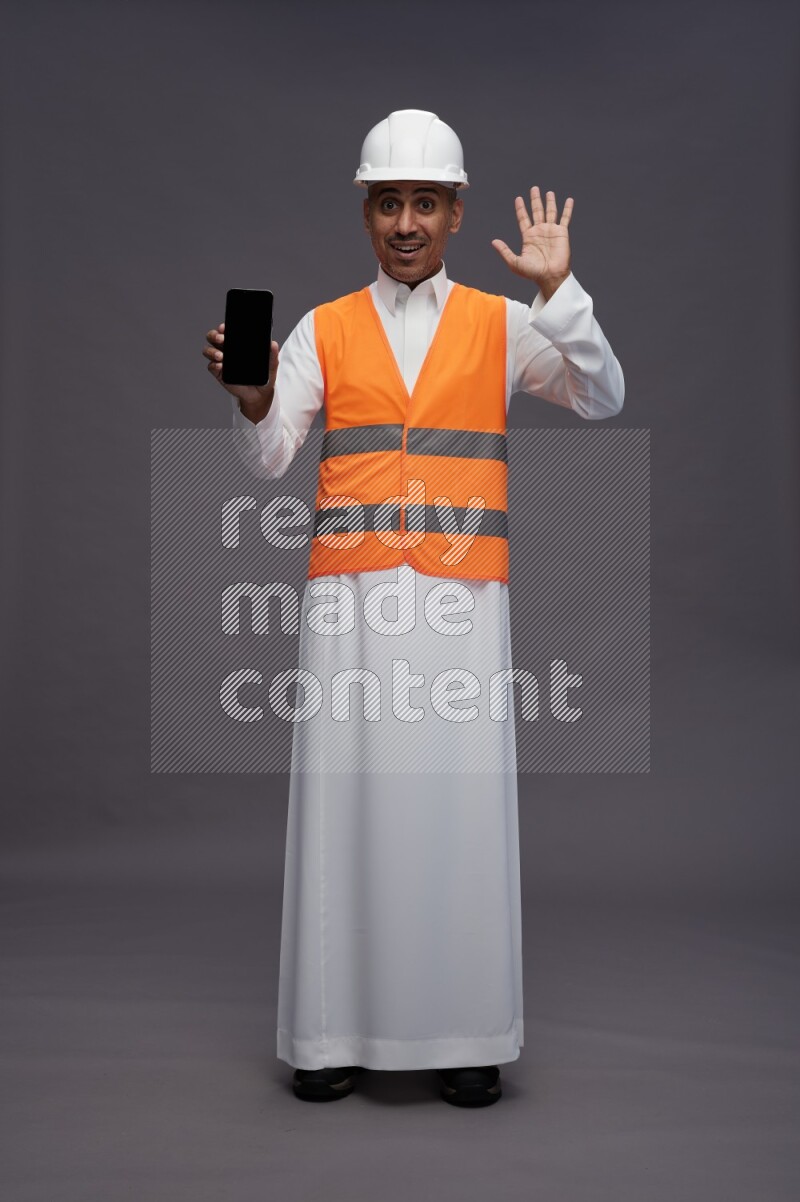 Saudi man wearing thob with engineer vest standing showing phone to camera on gray background