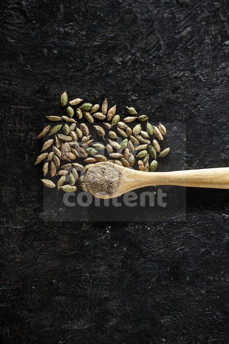 A wooden spoon full of cardamom powder and cardamom seeds next of it on textured black flooring