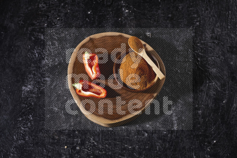 A wooden bowl full of ground paprika powder and sliced red bell pepper beside it, all on a wooden tray on black background