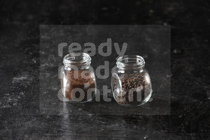 Glass spice jars full of cloves and cloves powder on textured black flooring