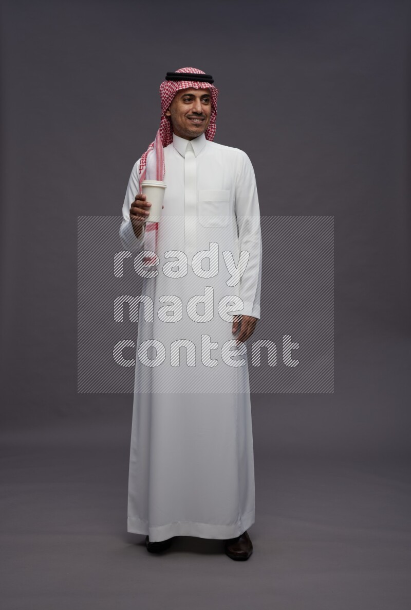 Saudi man wearing thob and shomag standing holding paper cup on gray background