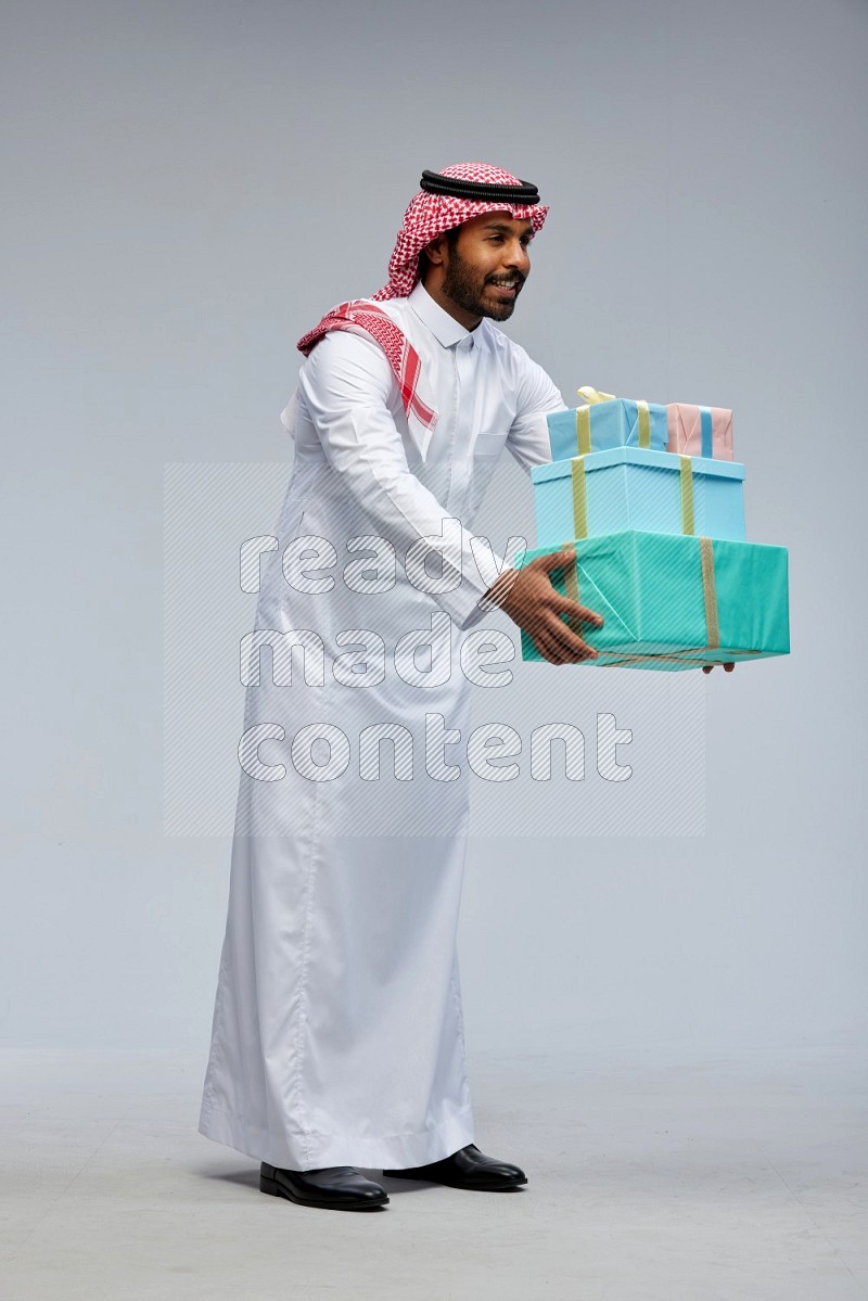 Saudi man Wearing Thob and shomag standing holding gift box on Gray background