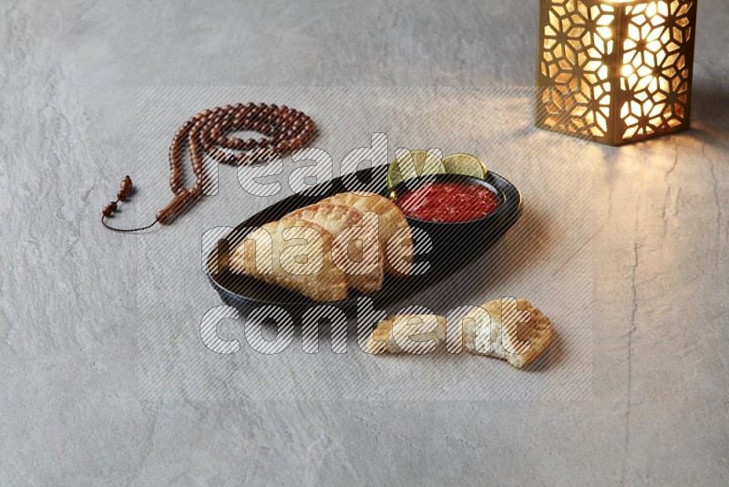 Three fried sambosas in an oval shaped black plate and a red sauce in a black round ramekin with a brown misbaha and a golden lantern on the side on a gray background