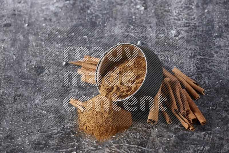 Black pottery bowl over filled with cinnamon powder and cinnamon sticks around the bowl on a textured black background