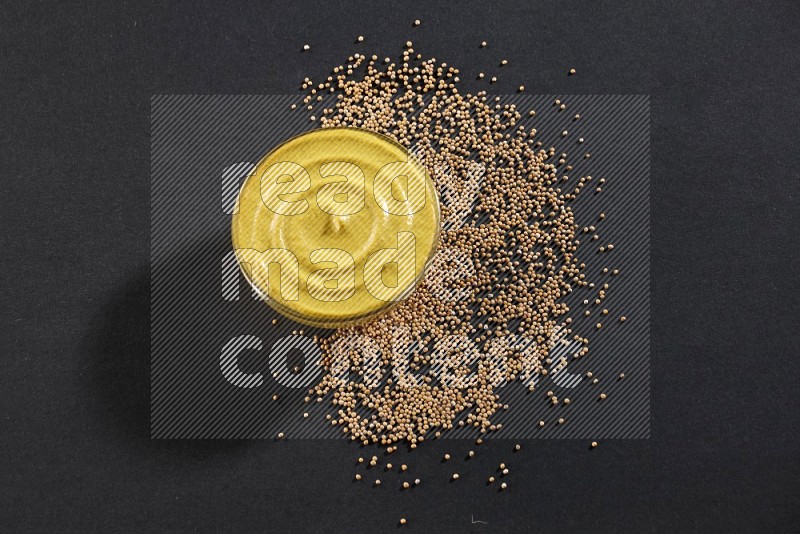A glass bowl full of mustard paste with mustard seeds underneath on black flooring in different angles