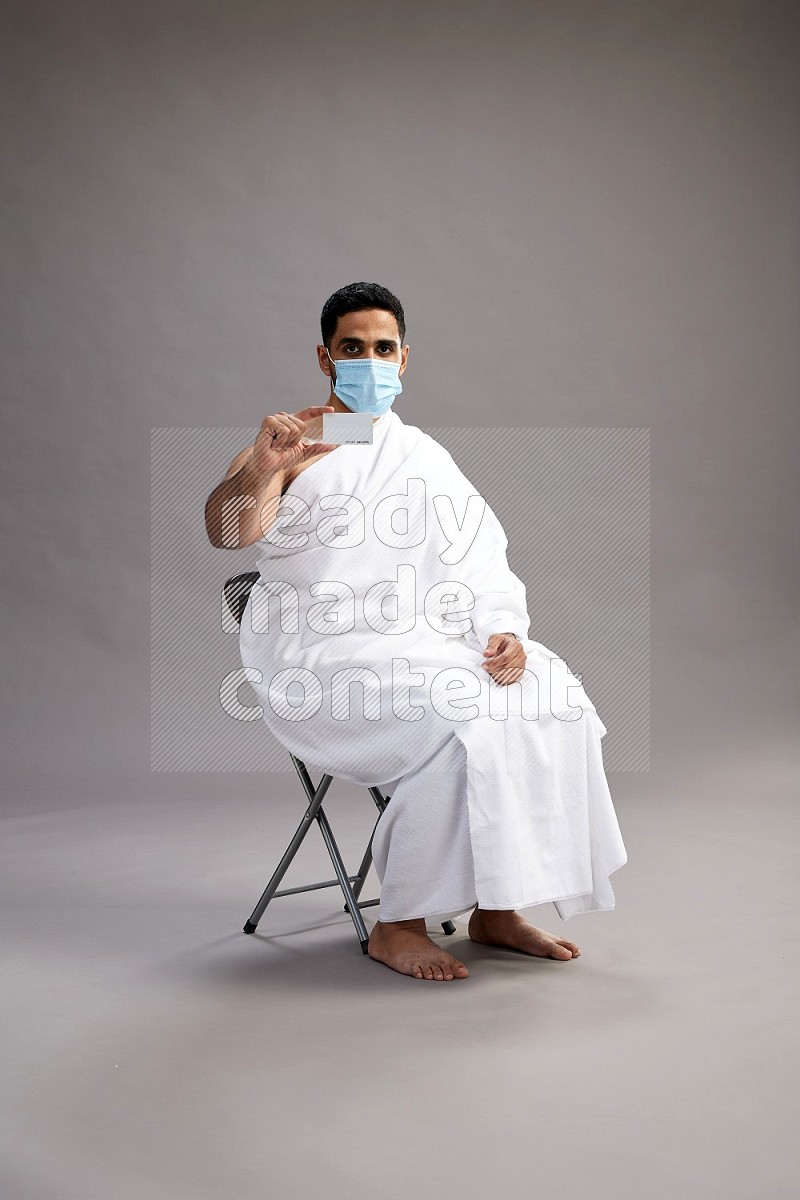 A man wearing Ehram with face mask sitting on chair holding ATM card on gray background