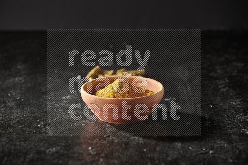 A wooden bowl full of turmeric powder with dried turmeric fingers on textured black flooring