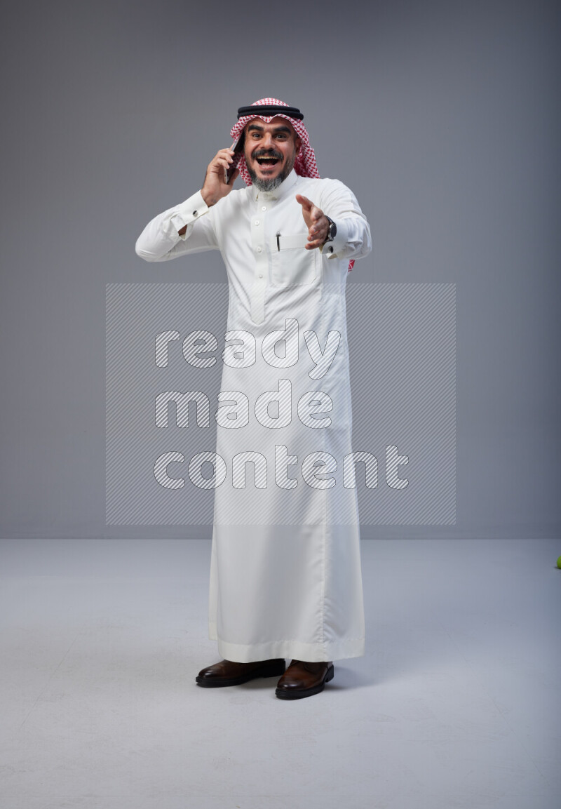 Saudi man Wearing Thob and red Shomag standing talking on phone on Gray background