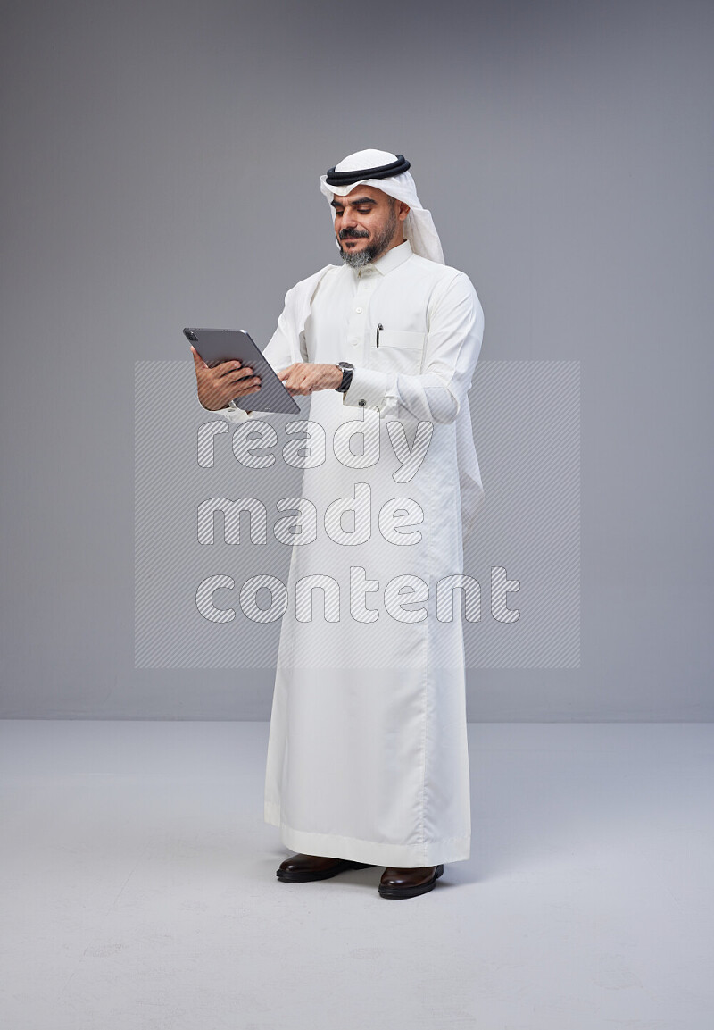 Saudi man Wearing Thob and white Shomag standing working on tablet on Gray background