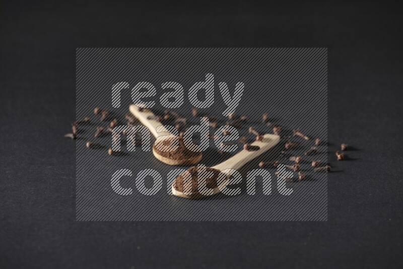 2 wooden spoons full of cloves powder with spreaded whole cloves on a black flooring