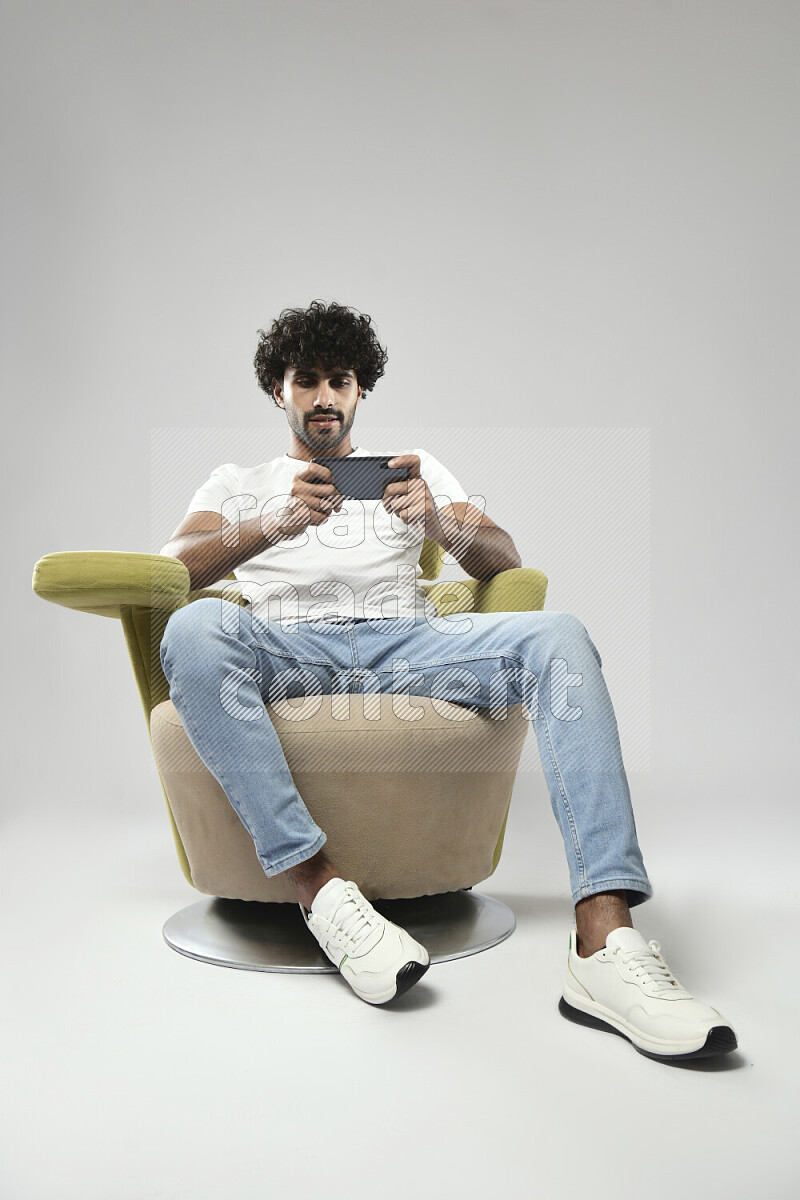 رجل يرتدي ملابس كاجوال يجلس علي كرسي و يلعب علي الهاتف علي خلفية بيضاء