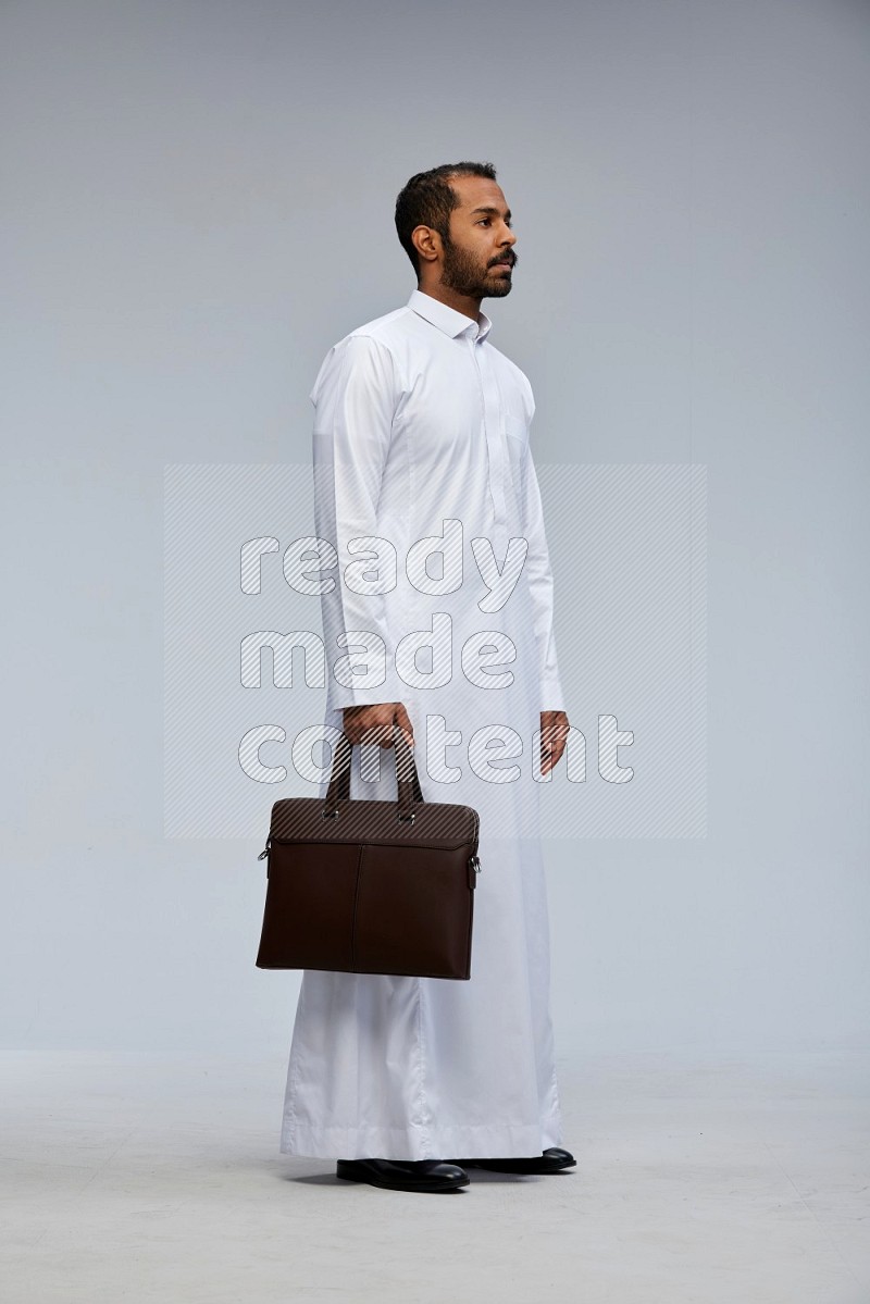 Saudi man wearing thob standing holding suitcase on gray background