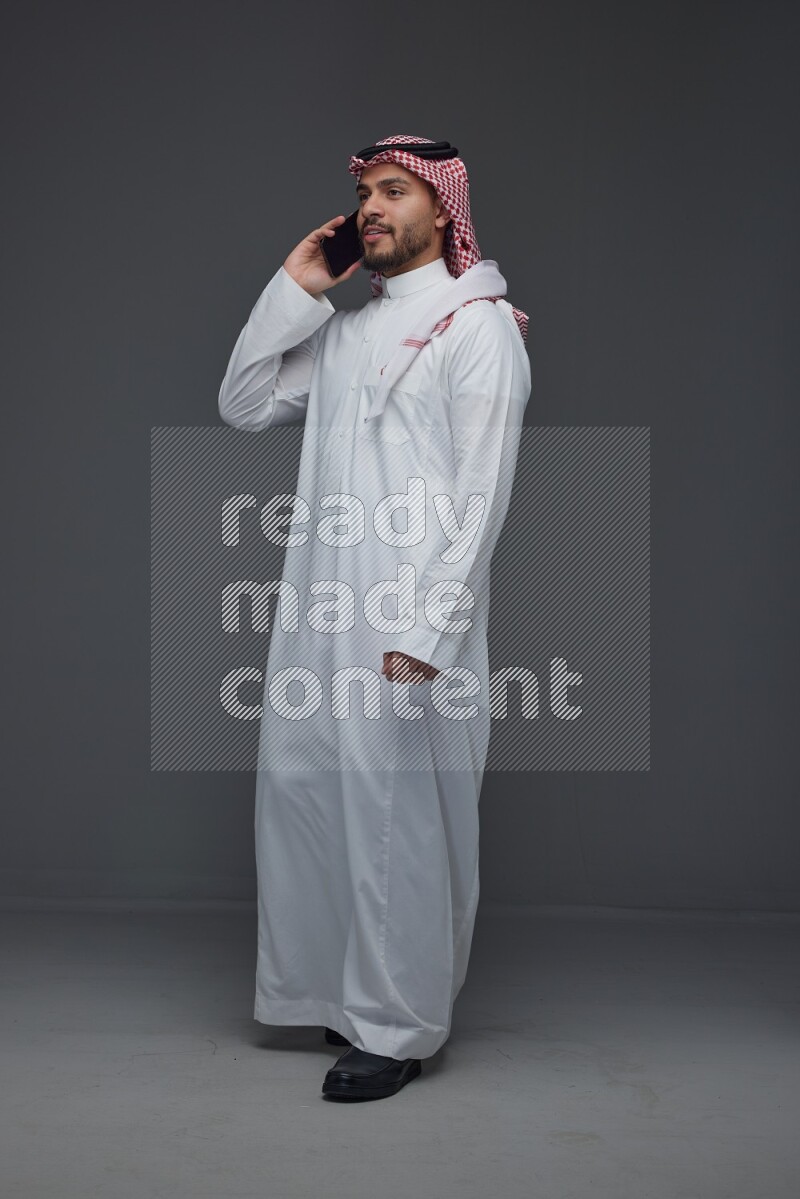 A Saudi man wearing Thobe and Shmagh talking in the phone eye level on a gray background