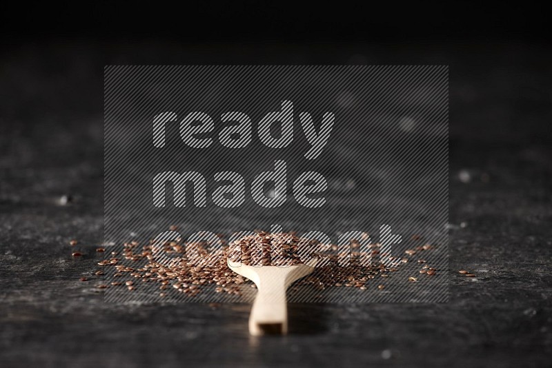 A wooden spoon full of flaxseeds and surrounded by seeds on a textured black flooring