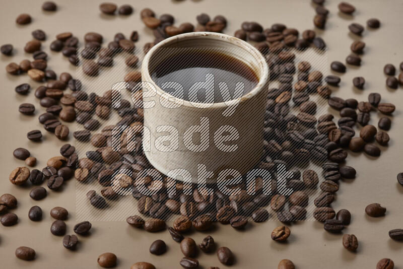 A beige pottery cup of coffee surrounded by roasted coffee beans on beige background