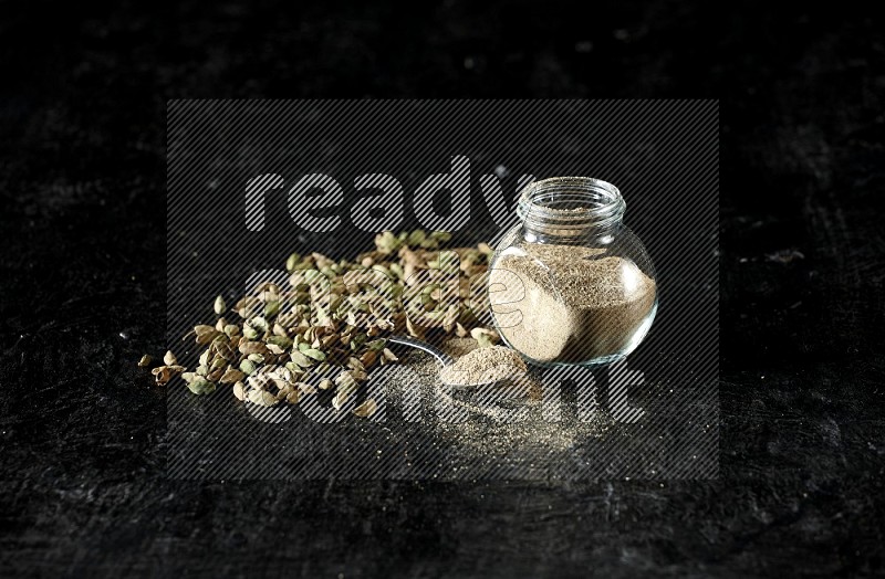 A glass spice jar and a metal spoon full of cardamom powder and cardamom seeds spreaded on textured black flooring