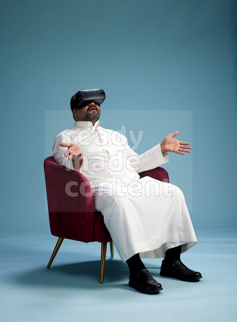 Saudi Man without shimag sitting on chair  gaming with VR on blue background