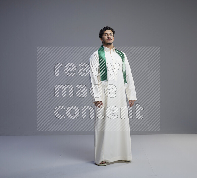 A Saudi man standing wearing thob and Saudi flag scarf on gray background