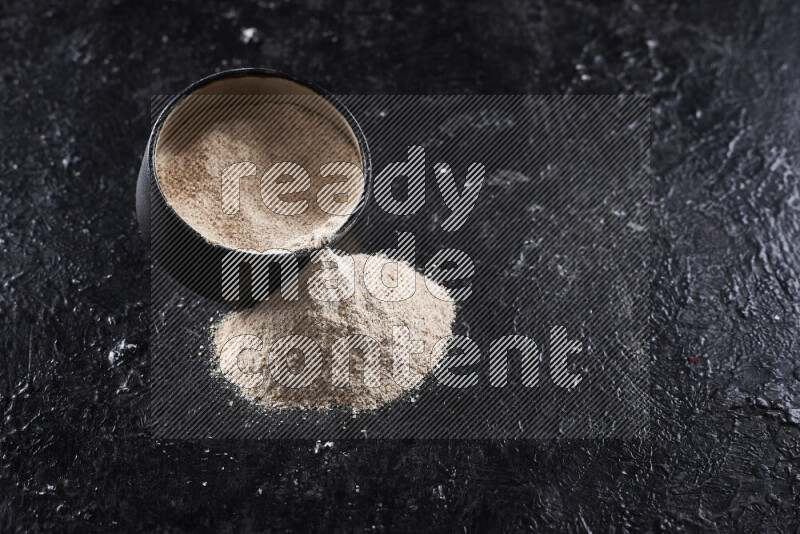 A black pottery bowl full of onion powder with fallen powder from it on black background