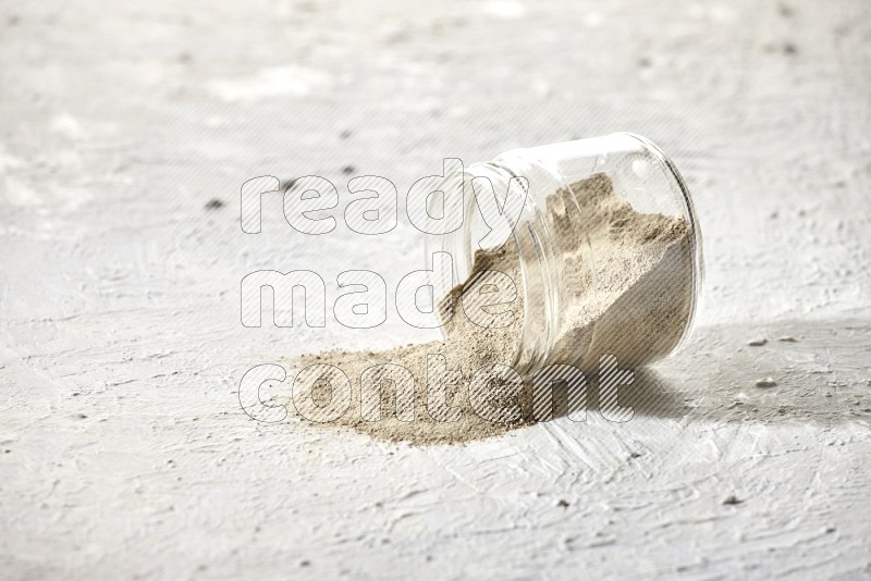 A flipped glass jar full of white pepper powder with spilled powder on textured white flooring