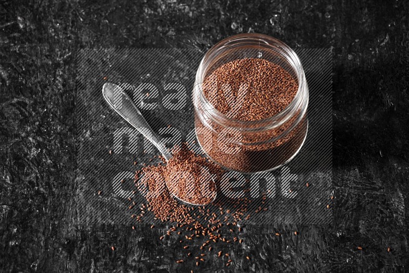 A glass jar full of garden cress seeds with a metal spoon full of the seeds on a textured black flooring